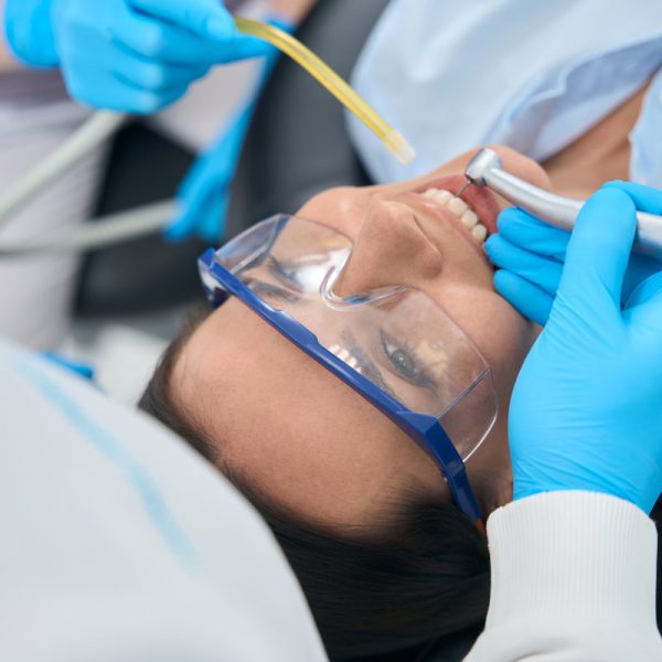 Stomatologist treating teeth to female client in protective glasses with electric drill that shooting water to her mouth, nurse holding suction device that removes saliva from mouth